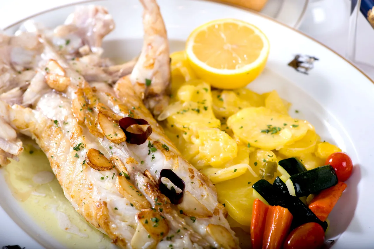 A grilled fish with garlic slices, potatoes, a lemon half and vegetables