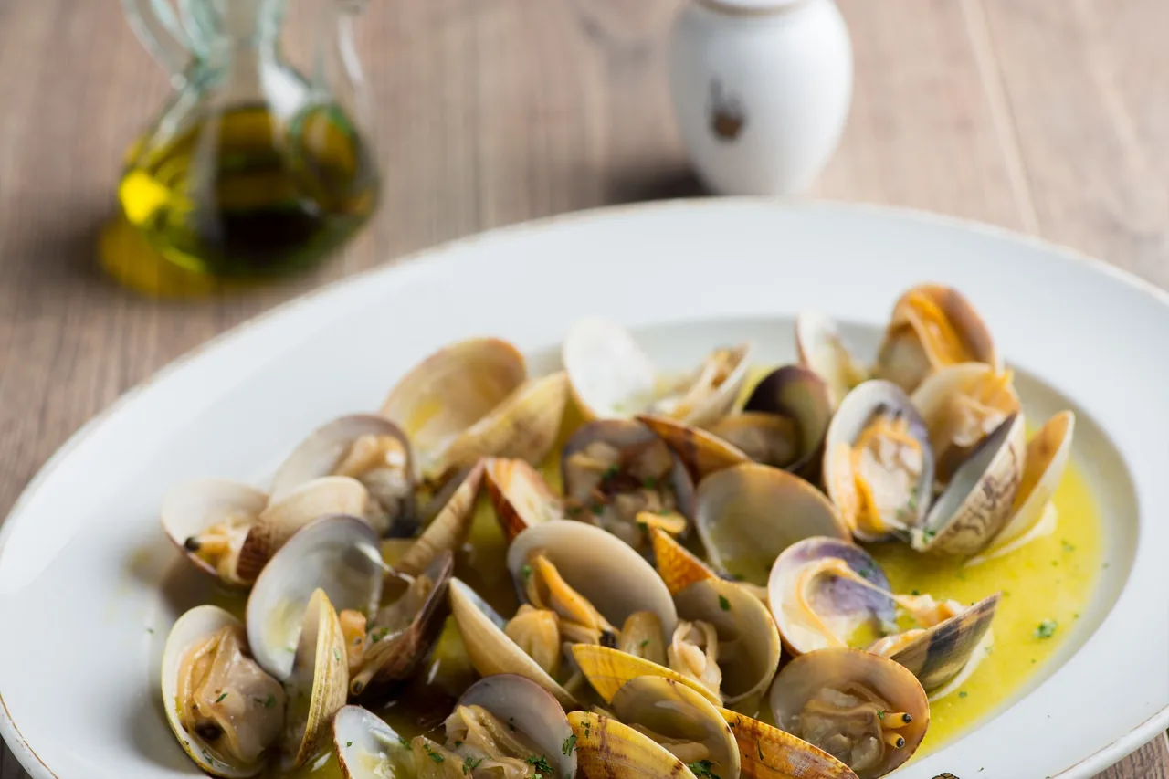Clams in a green sauce on a white plate beside a bottle of olive oil