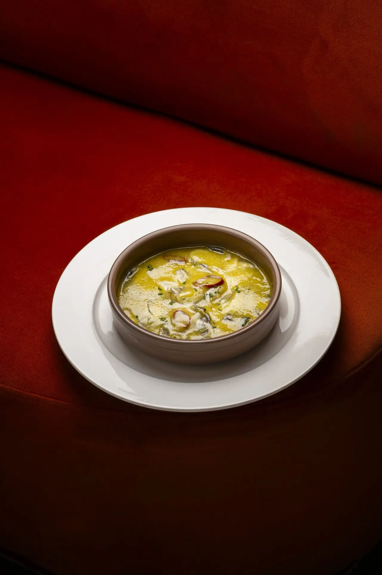 Earthenware bowl of a yellow egg-and-broth dish with sliced red peppers and chives, on a white plate over a red velvet banquette