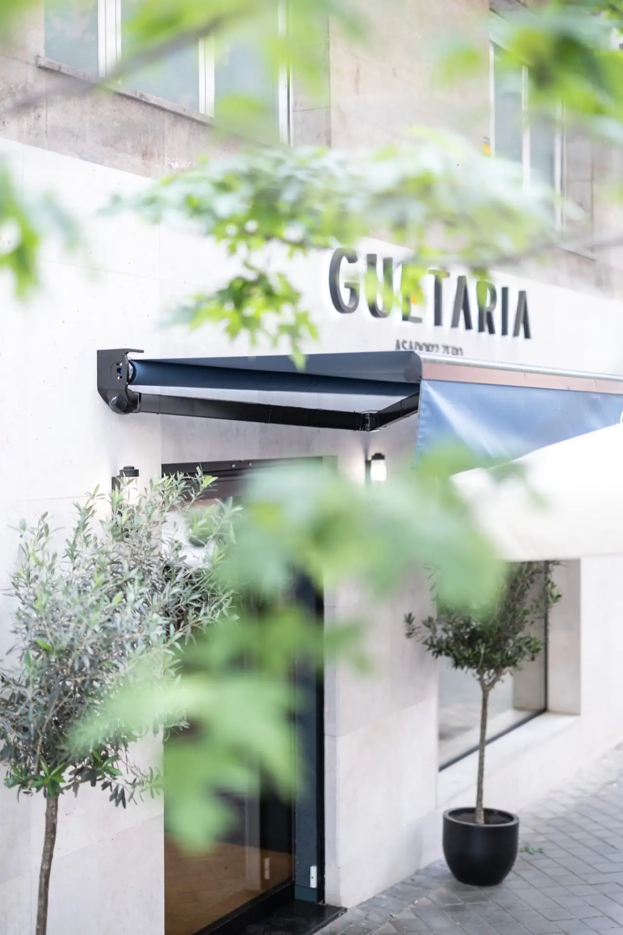 Stone-clad restaurant storefront with GUETARIA Asador signage, a dark retractable awning, doorway and potted olive trees, viewed through green foliage