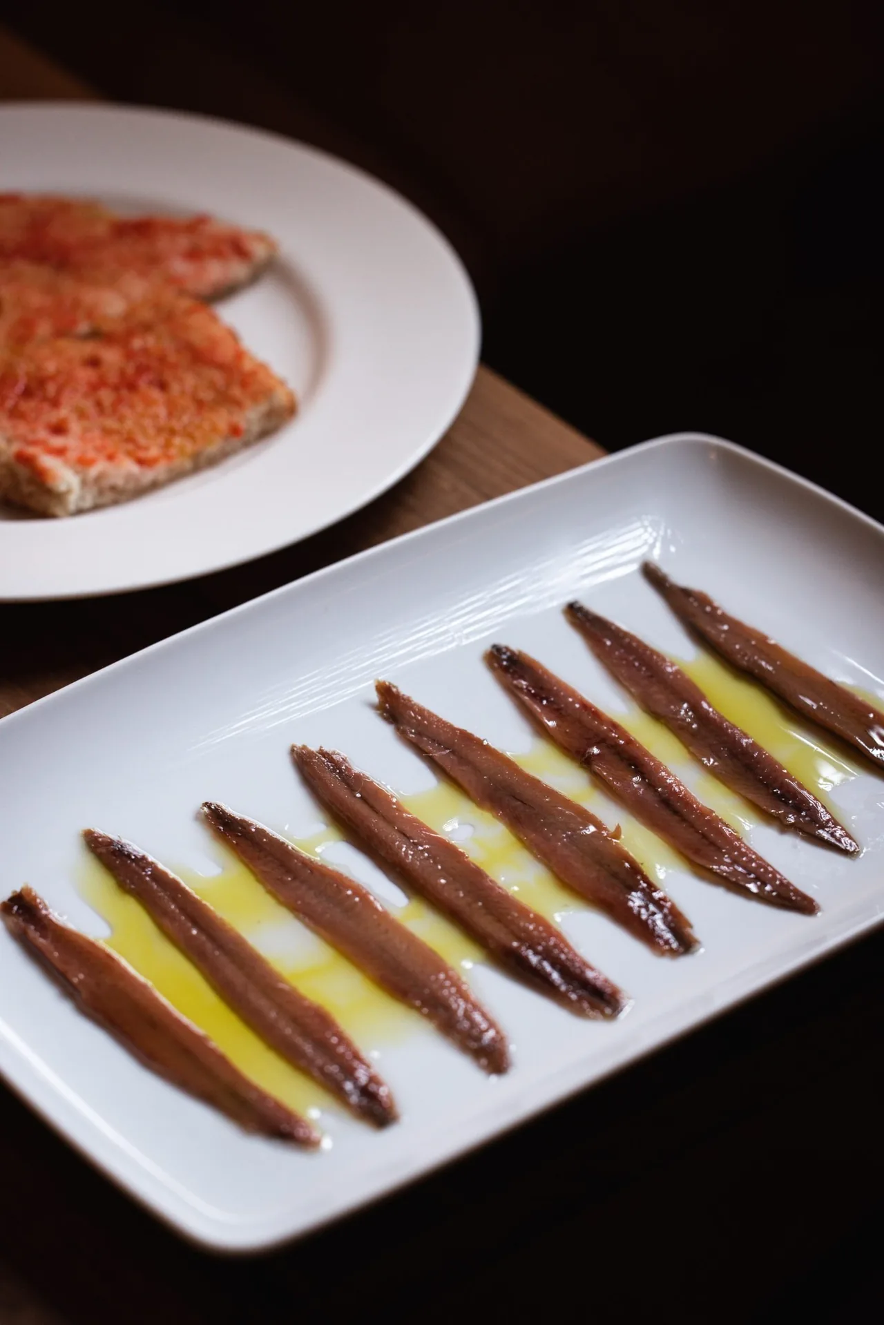 Anchovy fillets on a white rectangular plate beside a slice of tomato bread