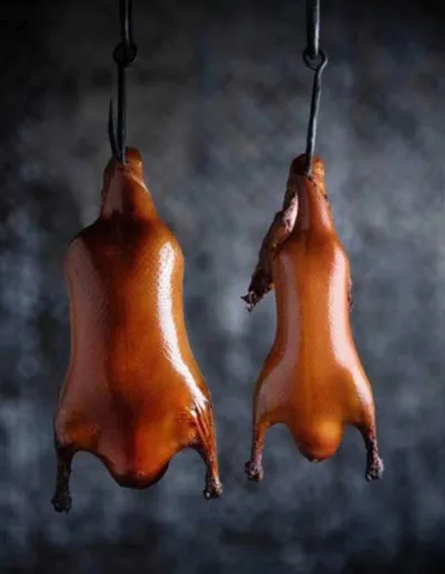 Two lacquered roast ducks hanging from iron hooks against a dark mottled backdrop, skin glossy and mahogany-brown