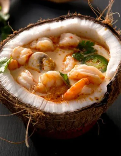 A coconut half filled with a pale coconut broth holding prawns, mushrooms, sliced green chilli and coriander leaves, set on dark slate