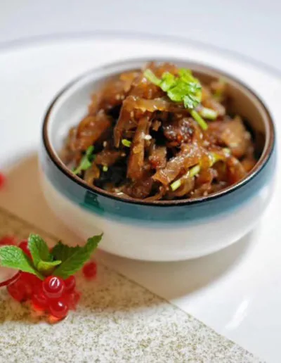 A small teal-rimmed ceramic bowl of glazed braised meat strips scattered with chopped spring onion, on a white plate beside a sprig of mint and a stem of redcurrants