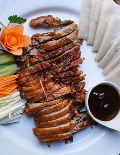 Sliced crisp-skinned roast duck fanned across a white plate with a stack of folded pancakes, a bowl of dark hoisin sauce, cucumber and spring onion batons and a carved carrot rose