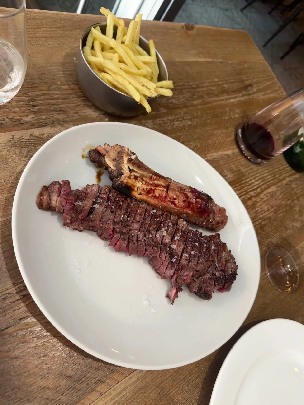 A grilled steak with a bone on a white plate with fries behind