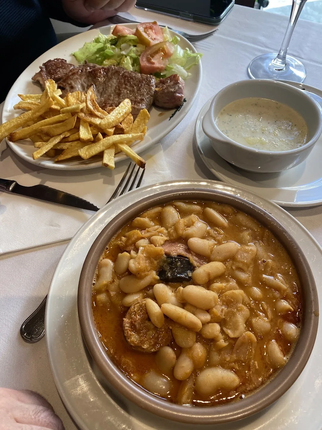A restaurant table with a bowl of white beans in orange broth with sausage, a plate of steak with fries and salad, and a bowl of white soup