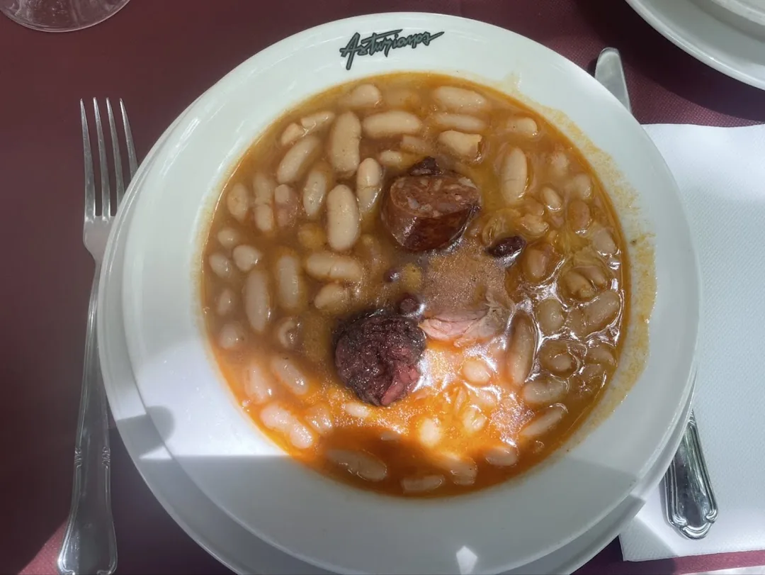 A close-up of white beans in orange broth with a slice of chorizo and a piece of black pudding