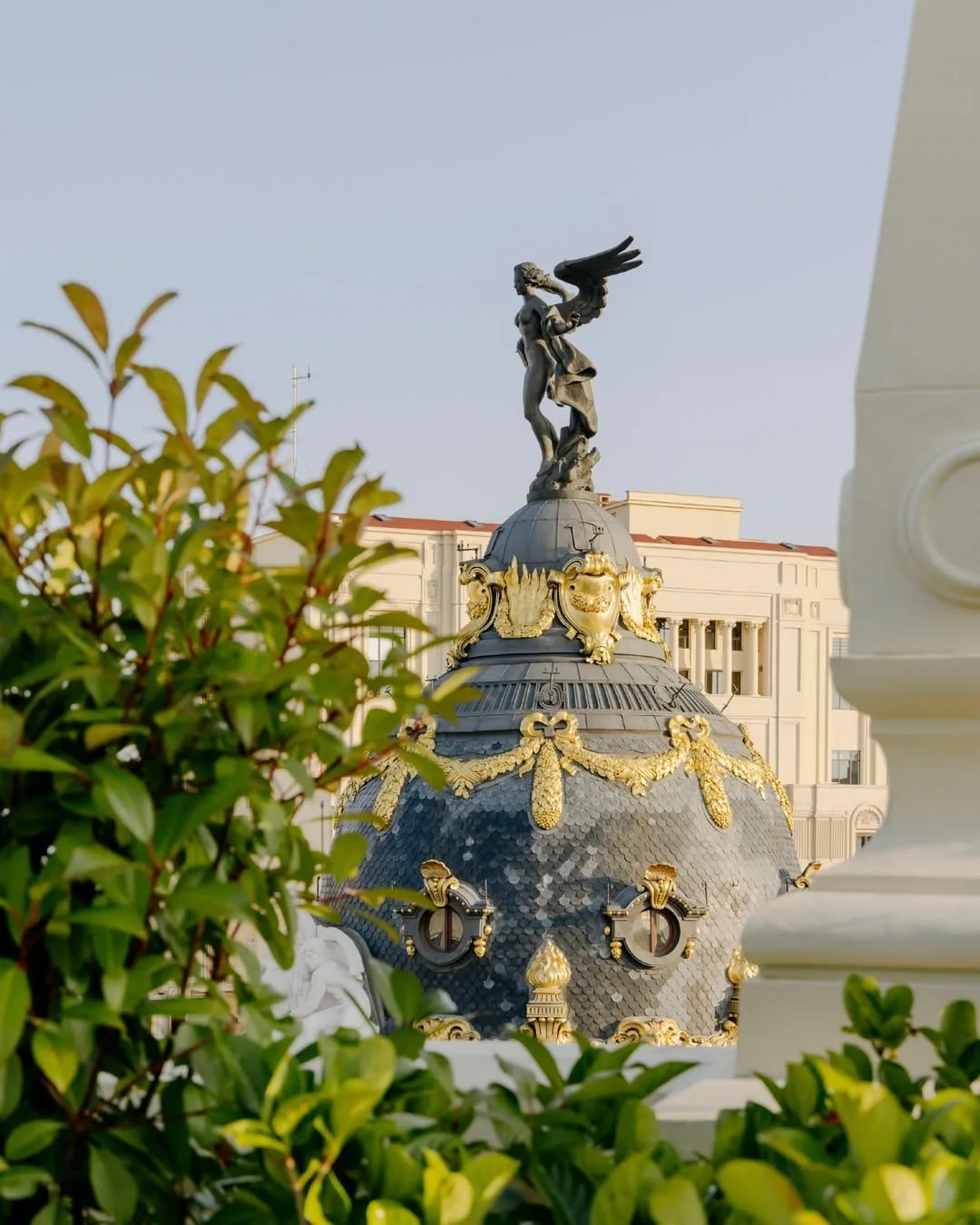 A dark domed roof with gold ornament and a winged statue seen through green leaves