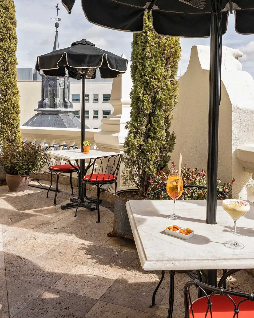 Rooftop terrace table with two cocktails and a small dish of fried snacks, city rooftops visible beyond the balustrade