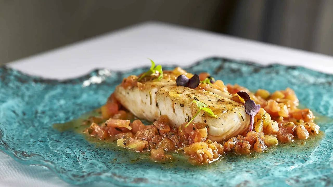 A fillet of white fish on chopped tomato with small leaves on a turquoise glass plate