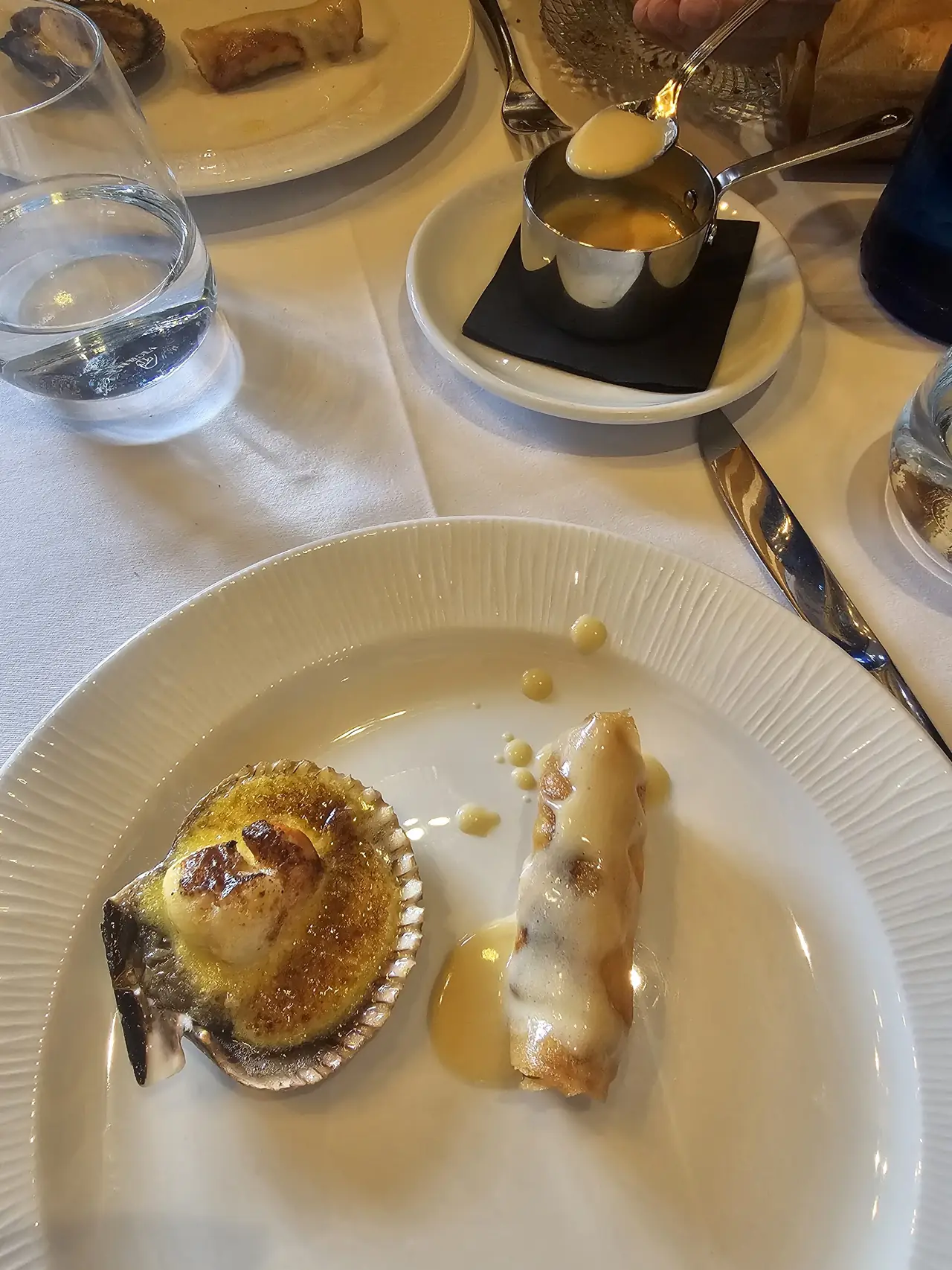 Gratinated scallop in its shell and a crepe roll with sauce, plated with a lit spoon flambeed in the background