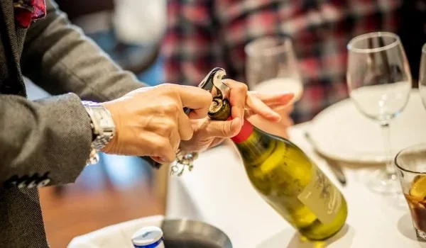 Hands drawing the foil from a bottle of white wine at a laid table, wine glasses and a poured glass just behind