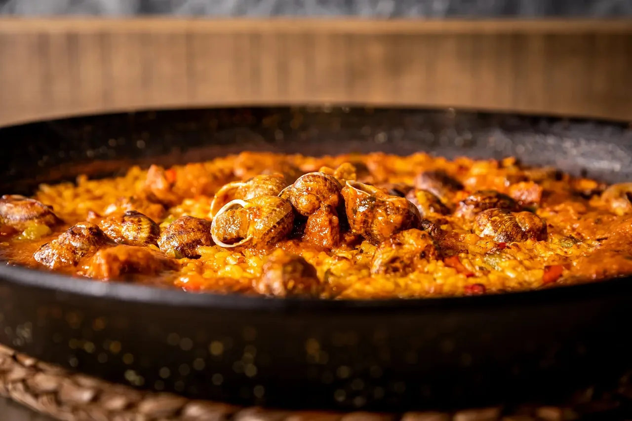 A wide steel pan of saffron-orange rice studded with whole snails in their shells and pieces of meat, steaming under kitchen light