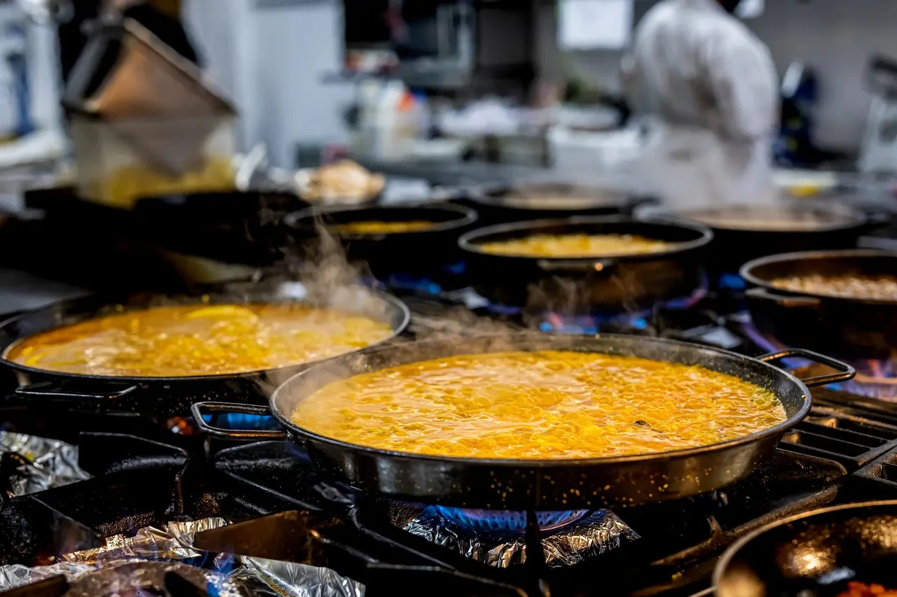 Several paella pans of bubbling saffron rice and stock over blue gas flames on a restaurant range, steam rising, a cook out of focus behind