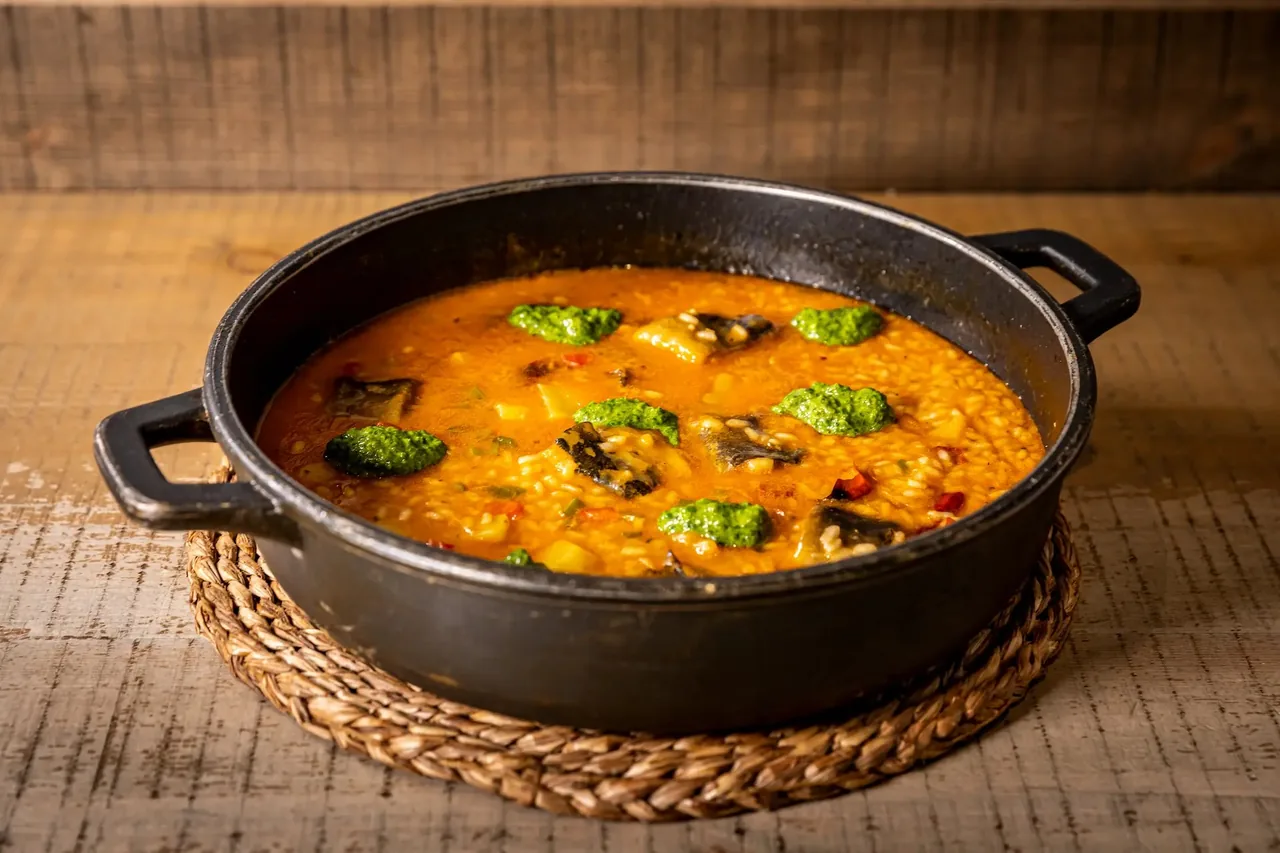 A black two-handled cast-iron dish of soupy orange rice with chunks of fish skin-side up and spoonfuls of bright green herb sauce, resting on a woven mat