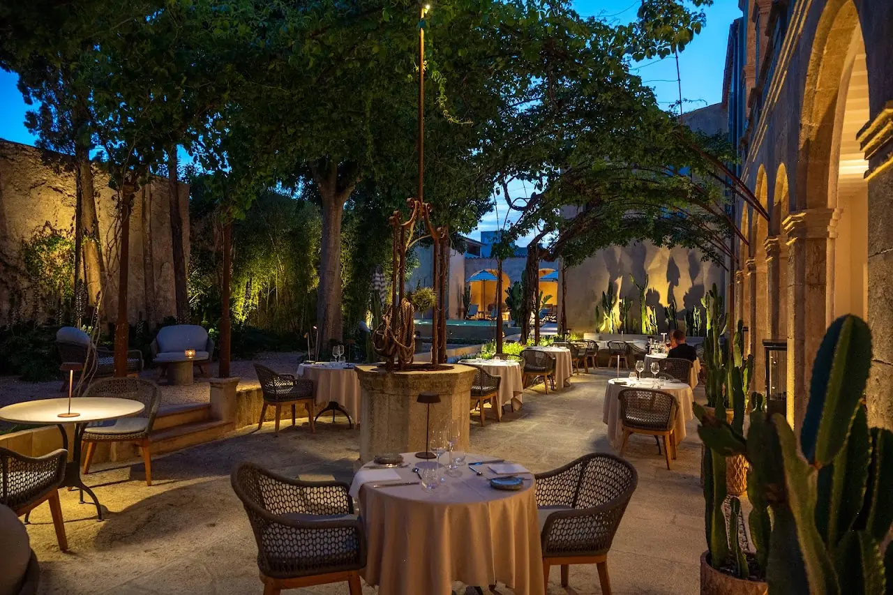 Candlelit courtyard dining area with set tables beneath trees and stone arches
