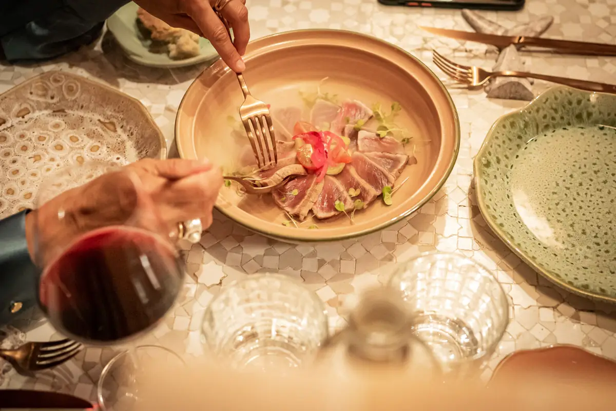Plate of sliced seared tuna with tomato and microgreens, a diner lifting a piece with a fork over a mosaic table set with red wine