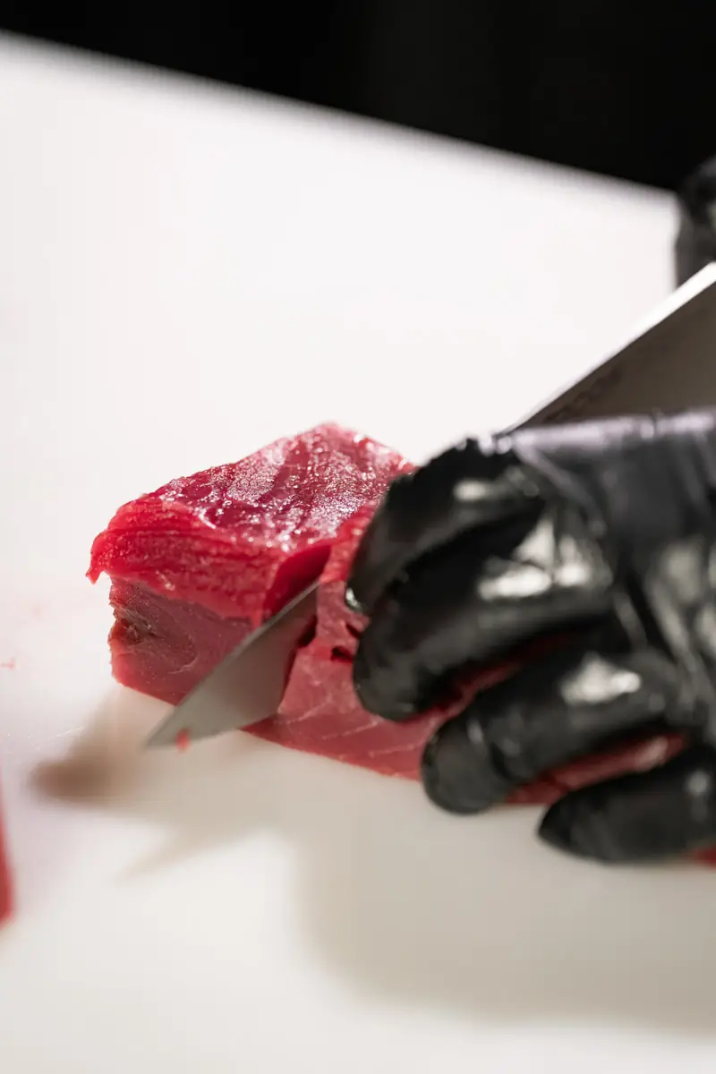 Black-gloved hands slicing a bright red raw tuna loin with a knife on a white cutting board