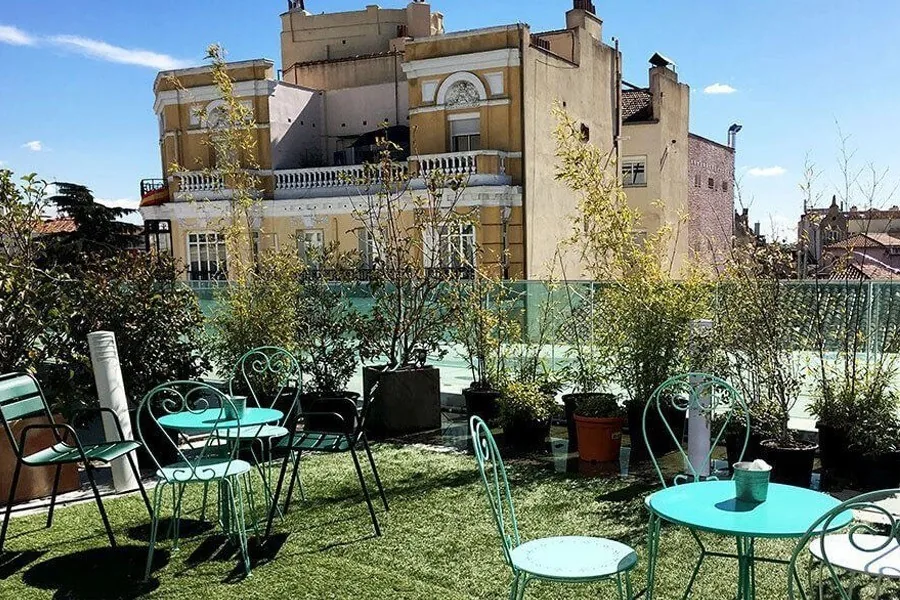 A rooftop terrace with turquoise metal chairs and tables on artificial grass beside potted plants and older buildings