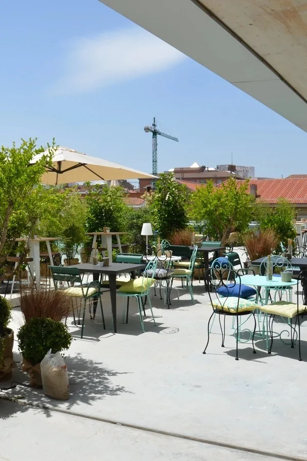 A sunny terrace with metal chairs, tables and umbrellas surrounded by plants