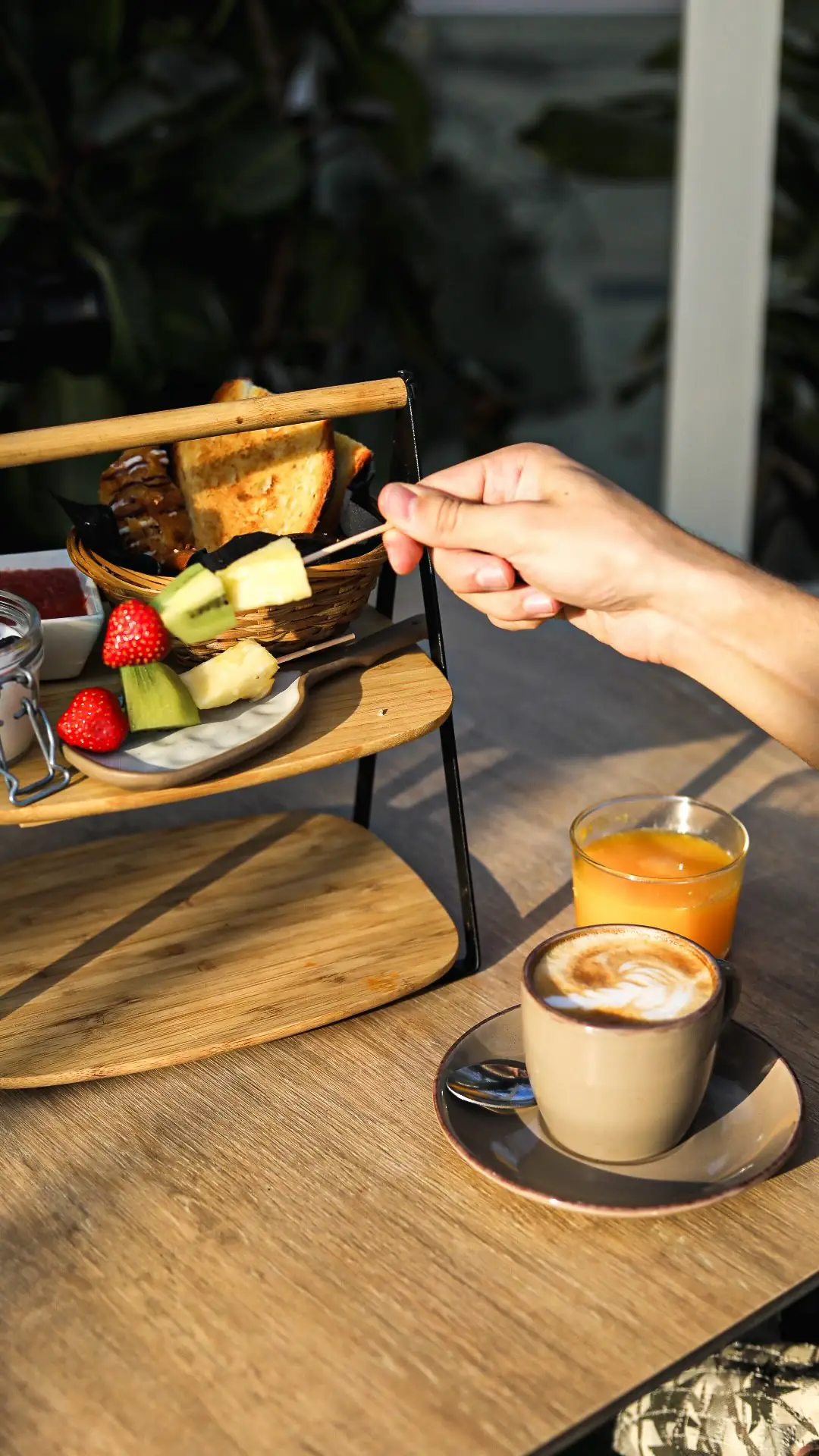 Three-tier wooden breakfast board with toast, croissant, and fruit skewer beside a latte and orange juice