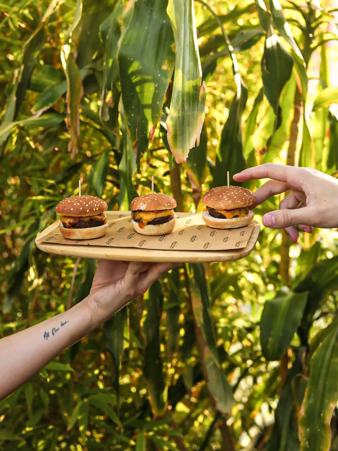 Three mini cheeseburgers with sesame buns served on a wooden board outdoors