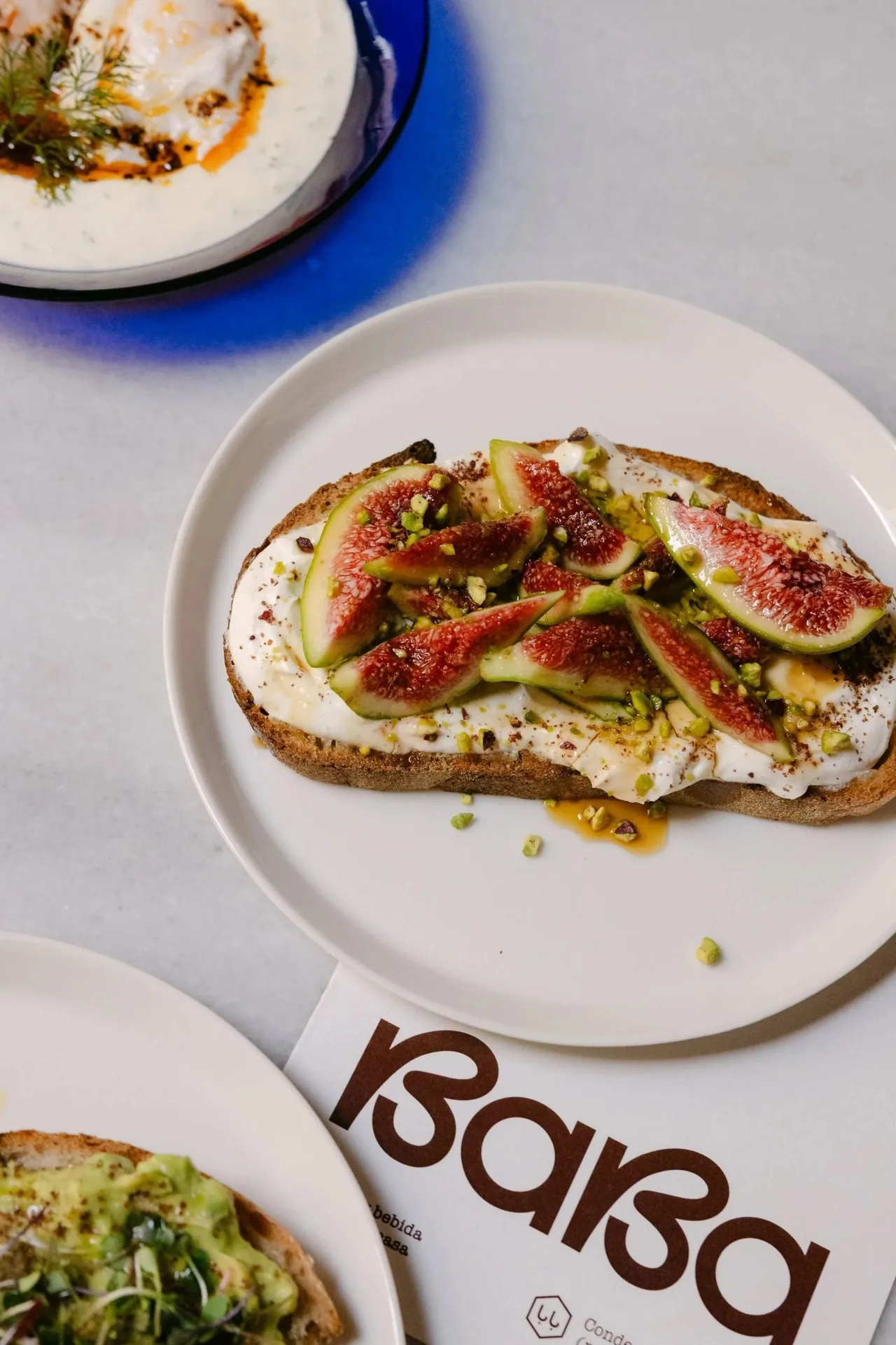 A slice of toast topped with white cream, fresh fig and chopped pistachios on a white plate