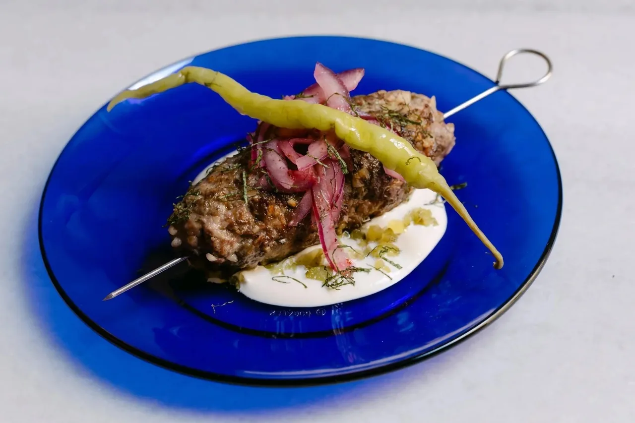 A grilled minced-meat skewer with pickled red onion and a green pepper on white sauce on a cobalt-blue glass plate