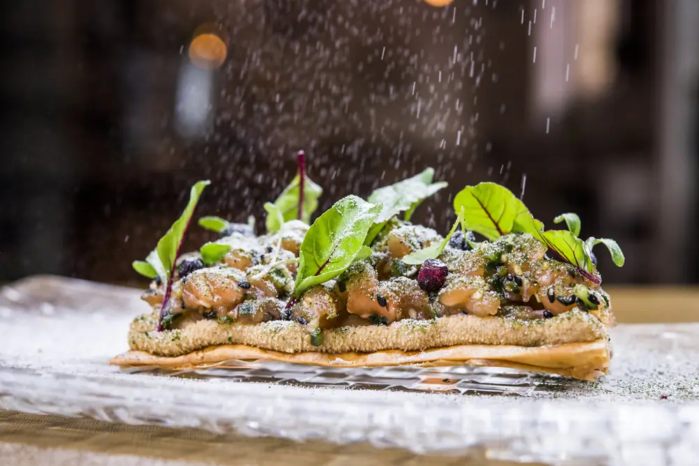 Crisp pastry base loaded with salmon tartare and red-veined leaves, being dusted with green powder from above.