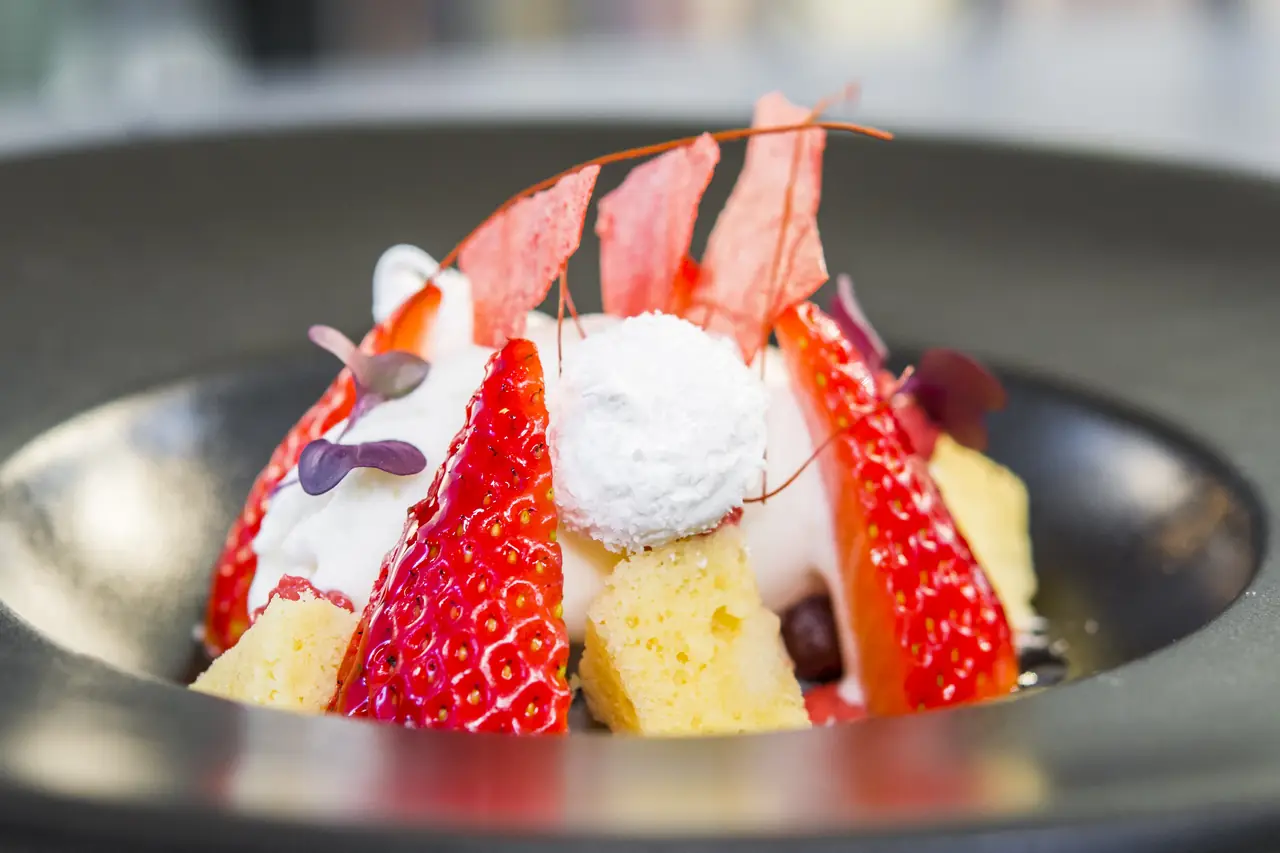 Dark bowl of strawberries with sponge cubes, a coconut-coated ball, cream and thin red tuiles.