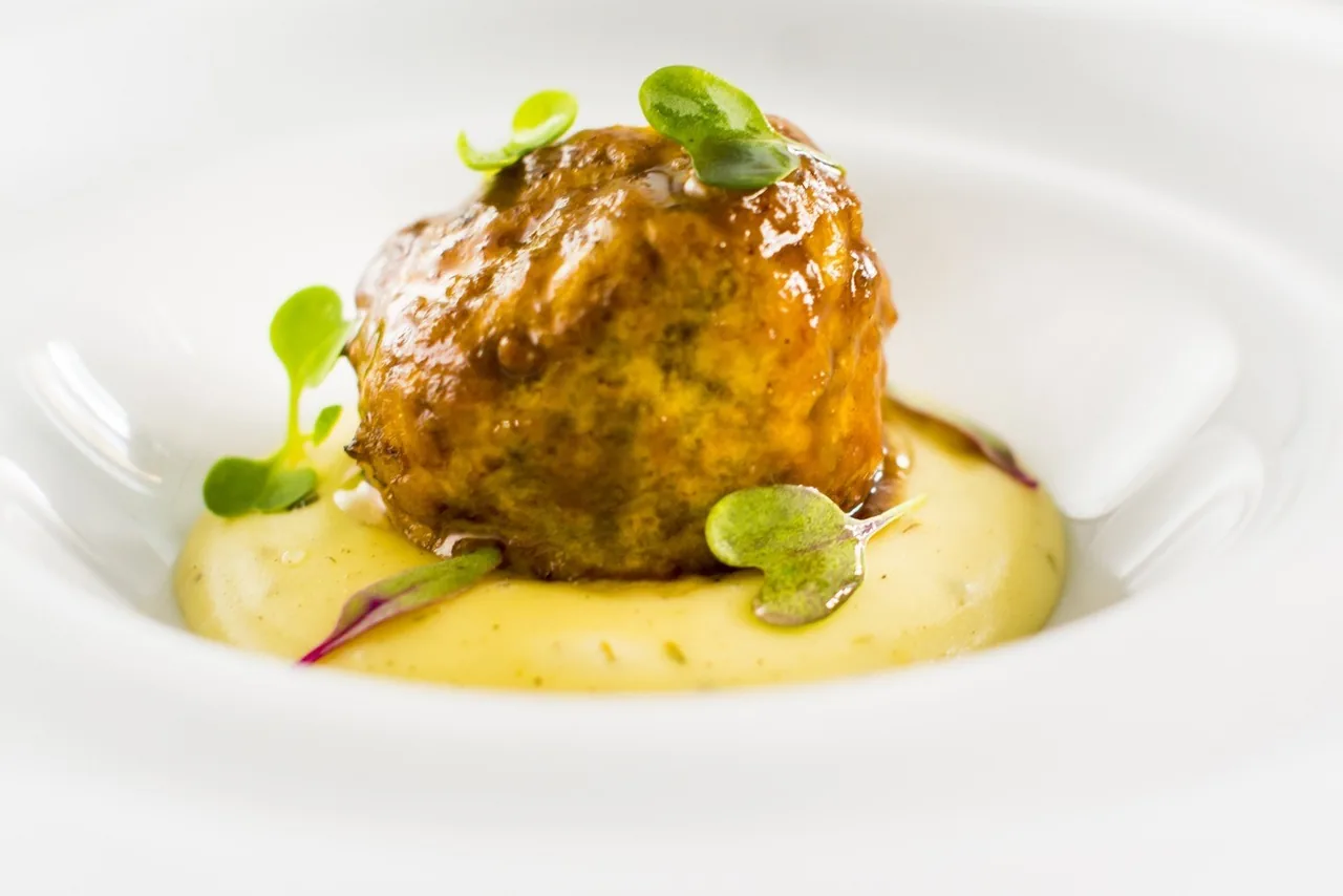 A round golden croquette on a yellow cream with a green garnish on a white plate