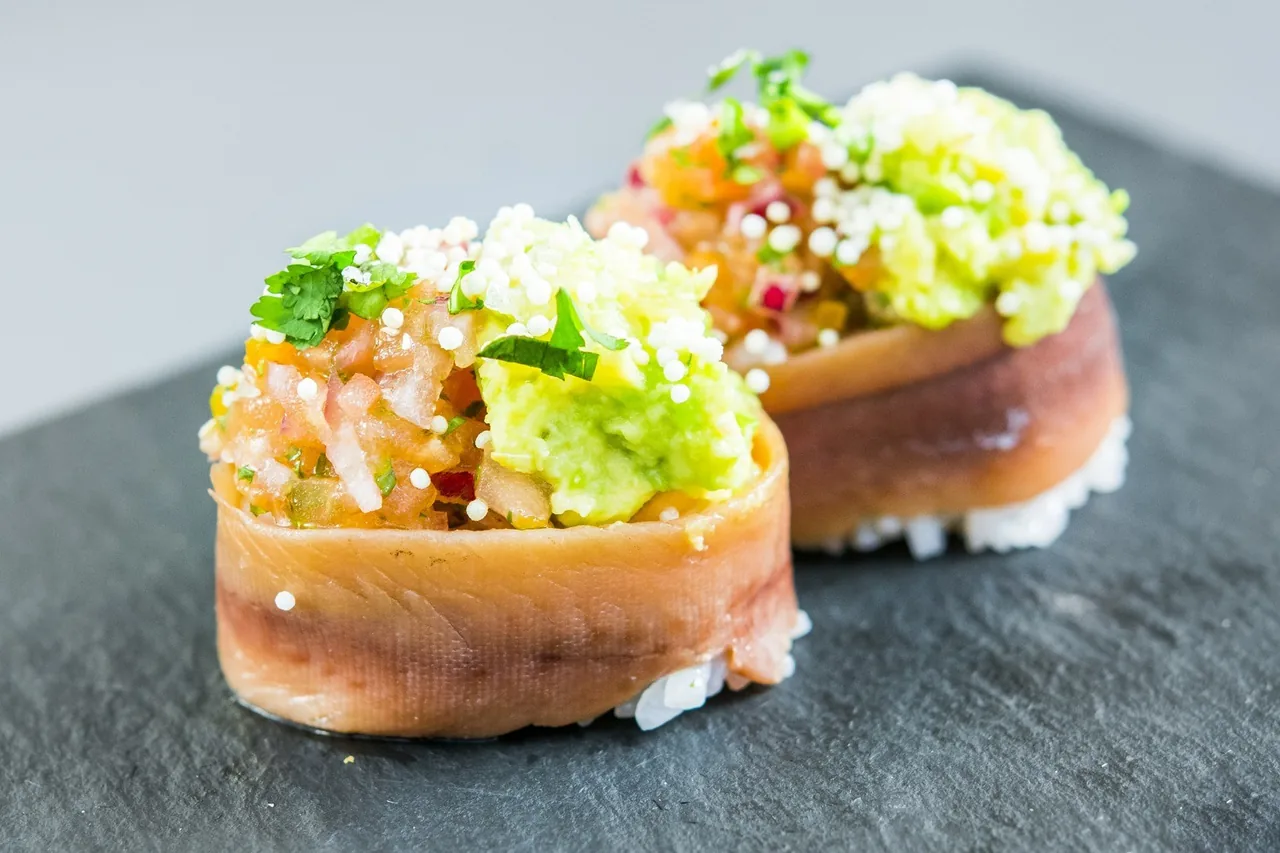 Two bites of raw fish topped with diced avocado and herbs on a slate board