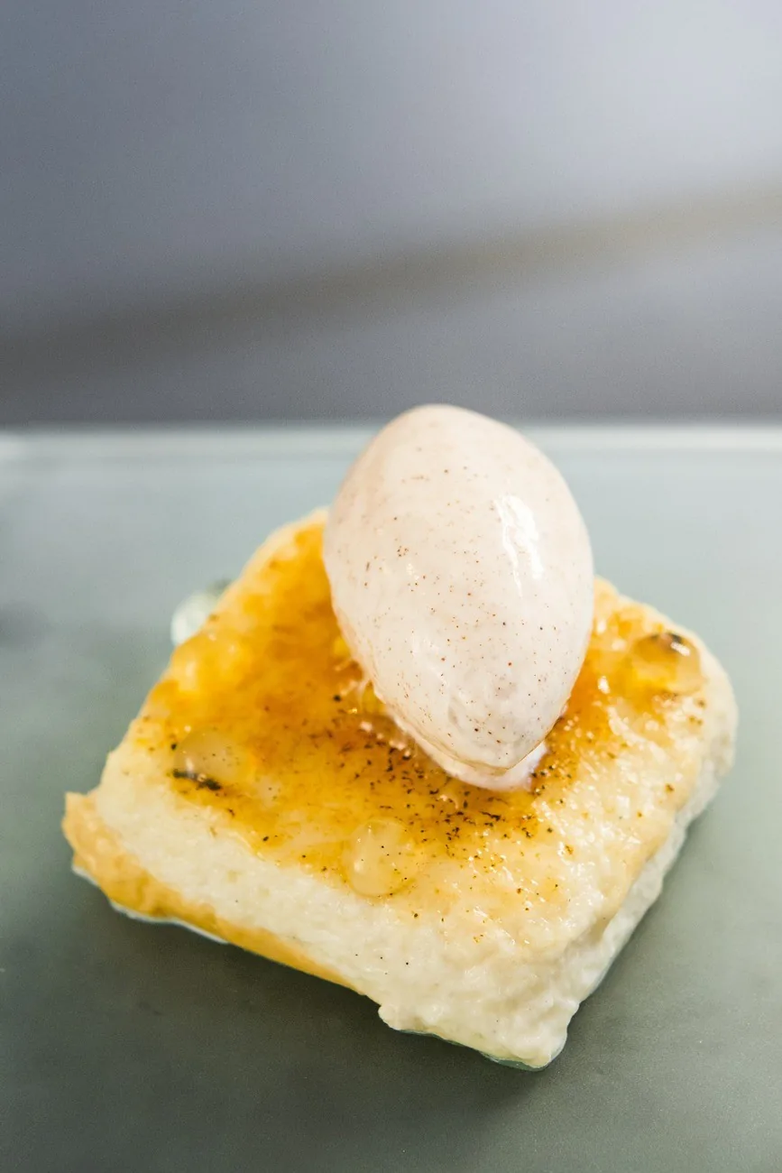 A square of golden caramelised bread topped with a quenelle of ice cream on a grey plate