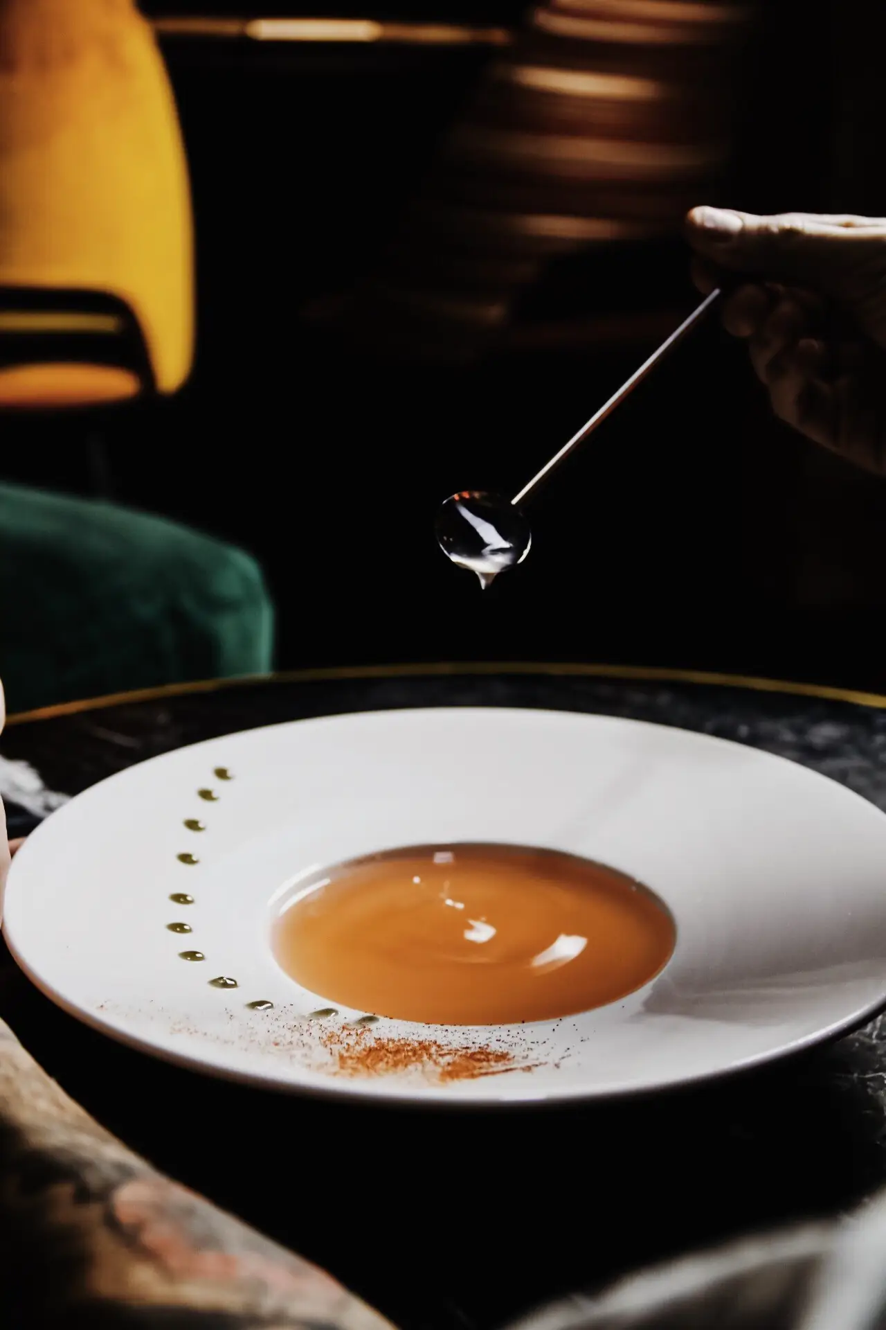 White plate with a pool of amber broth, a line of green oil drops and dusted spice, a hand adding a drop from a long spoon