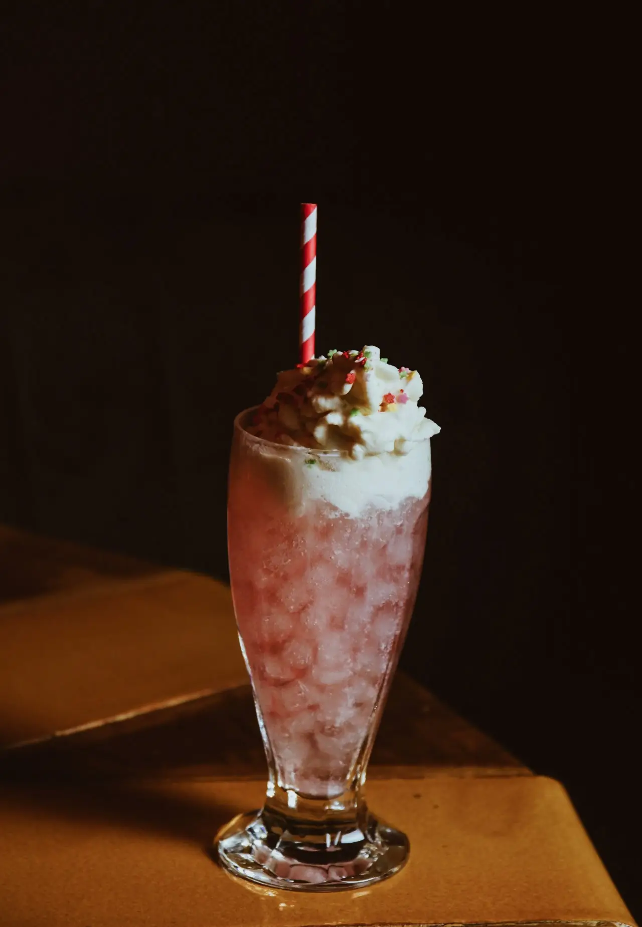 Tall glass of a pink iced drink topped with whipped cream, colored sprinkles and a red-and-white striped straw