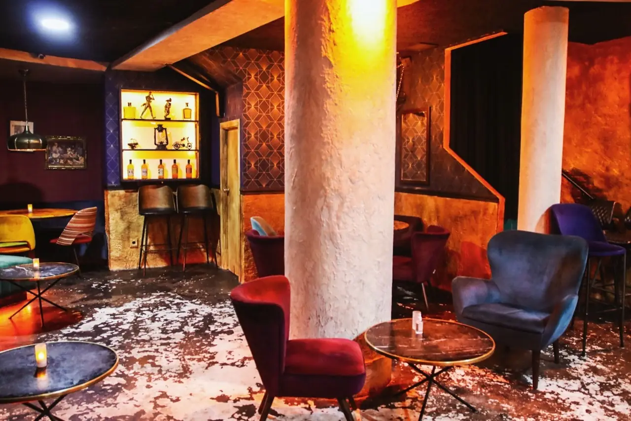 Dim lounge interior with velvet armchairs, marble bistro tables, a plaster column and a lit shelf of bottles and figurines