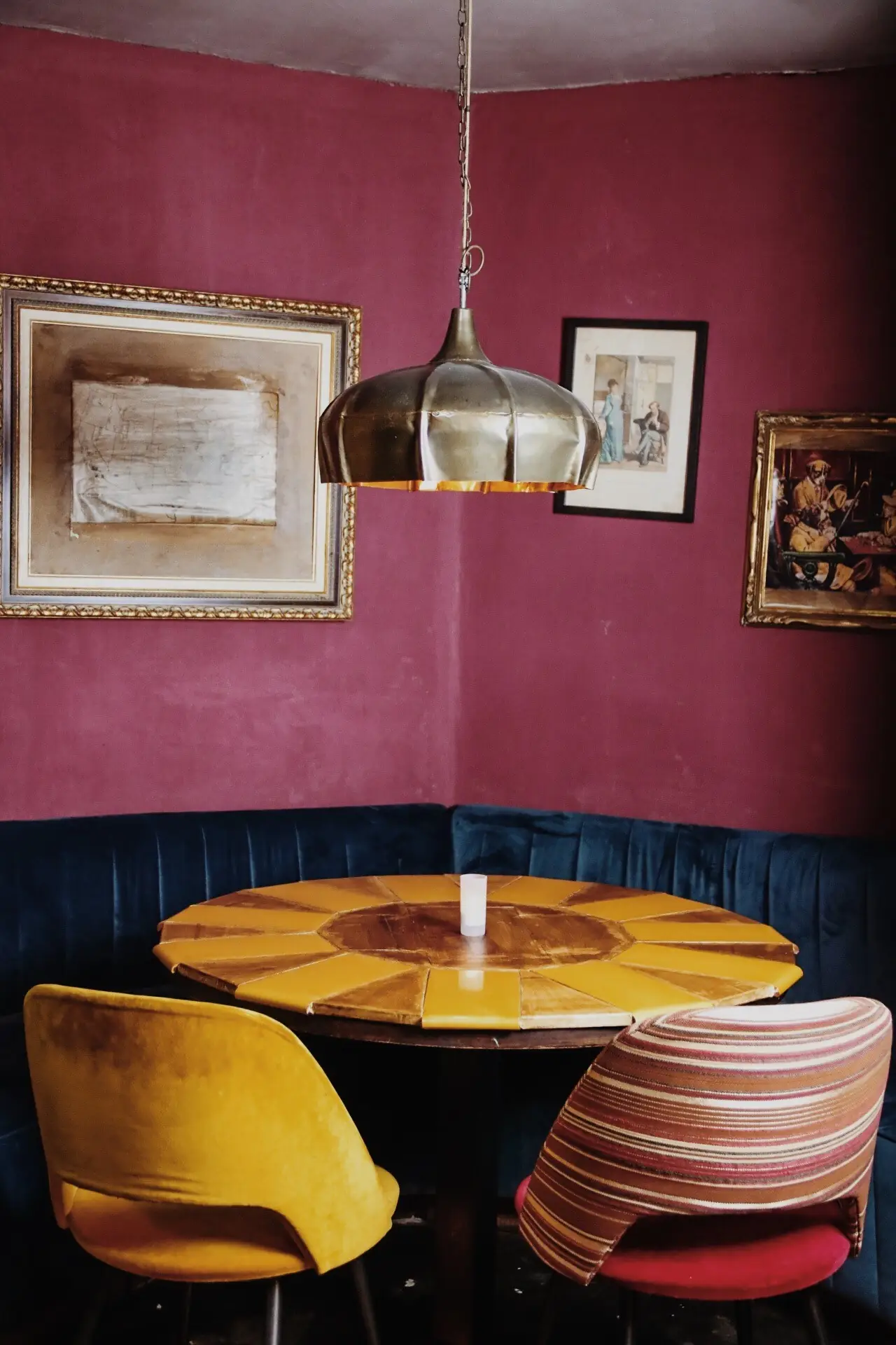 Corner booth with blue velvet banquette, a mustard-yellow segmented round table and framed art on a burgundy wall