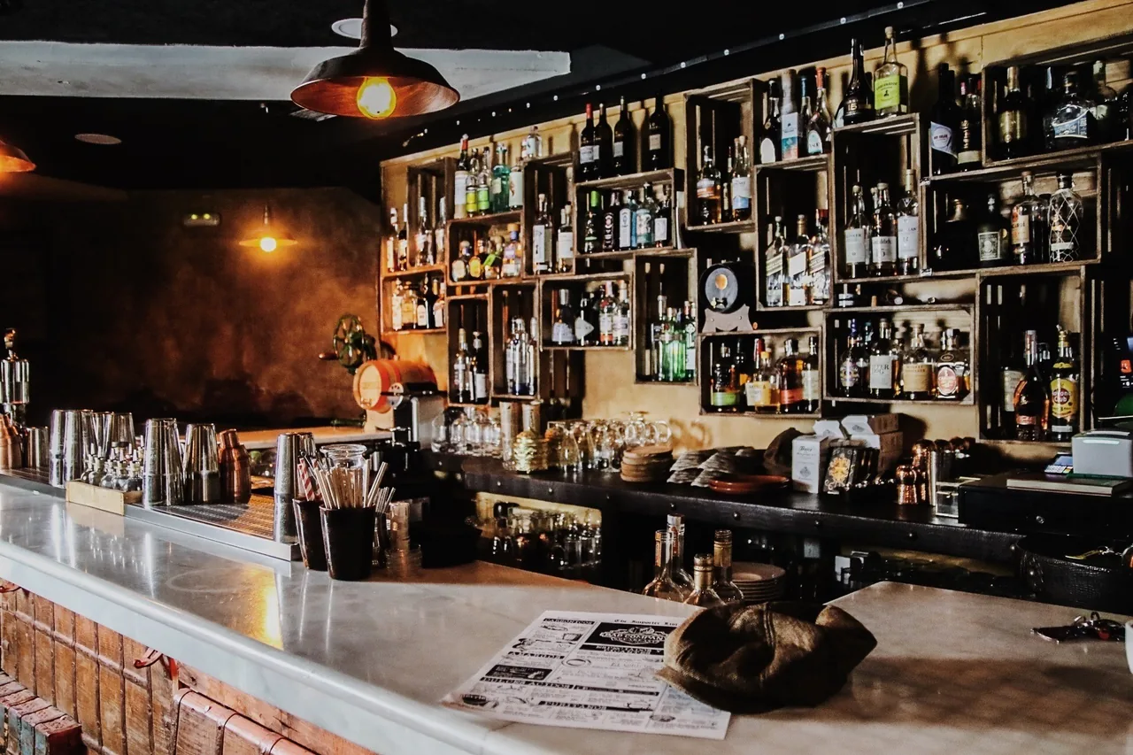 A marble bar counter with shakers and glasses in front of shelves of bottles in wooden crates