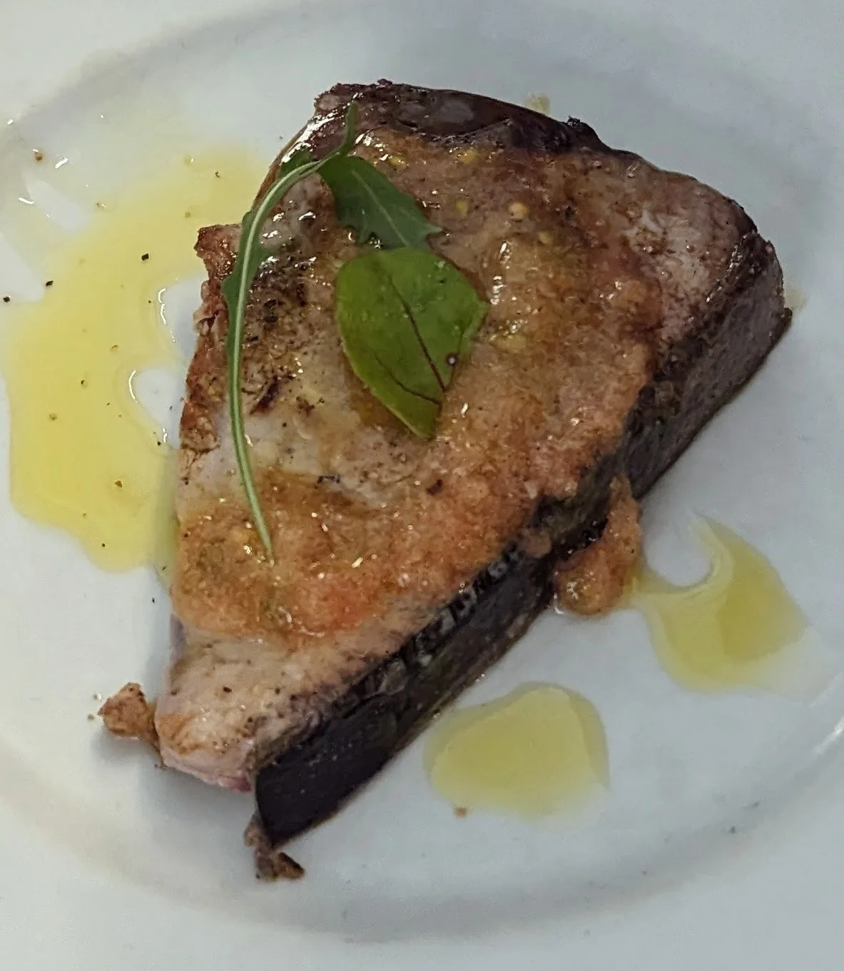 A thick grilled fish steak drizzled with oil and topped with a basil leaf on a white plate