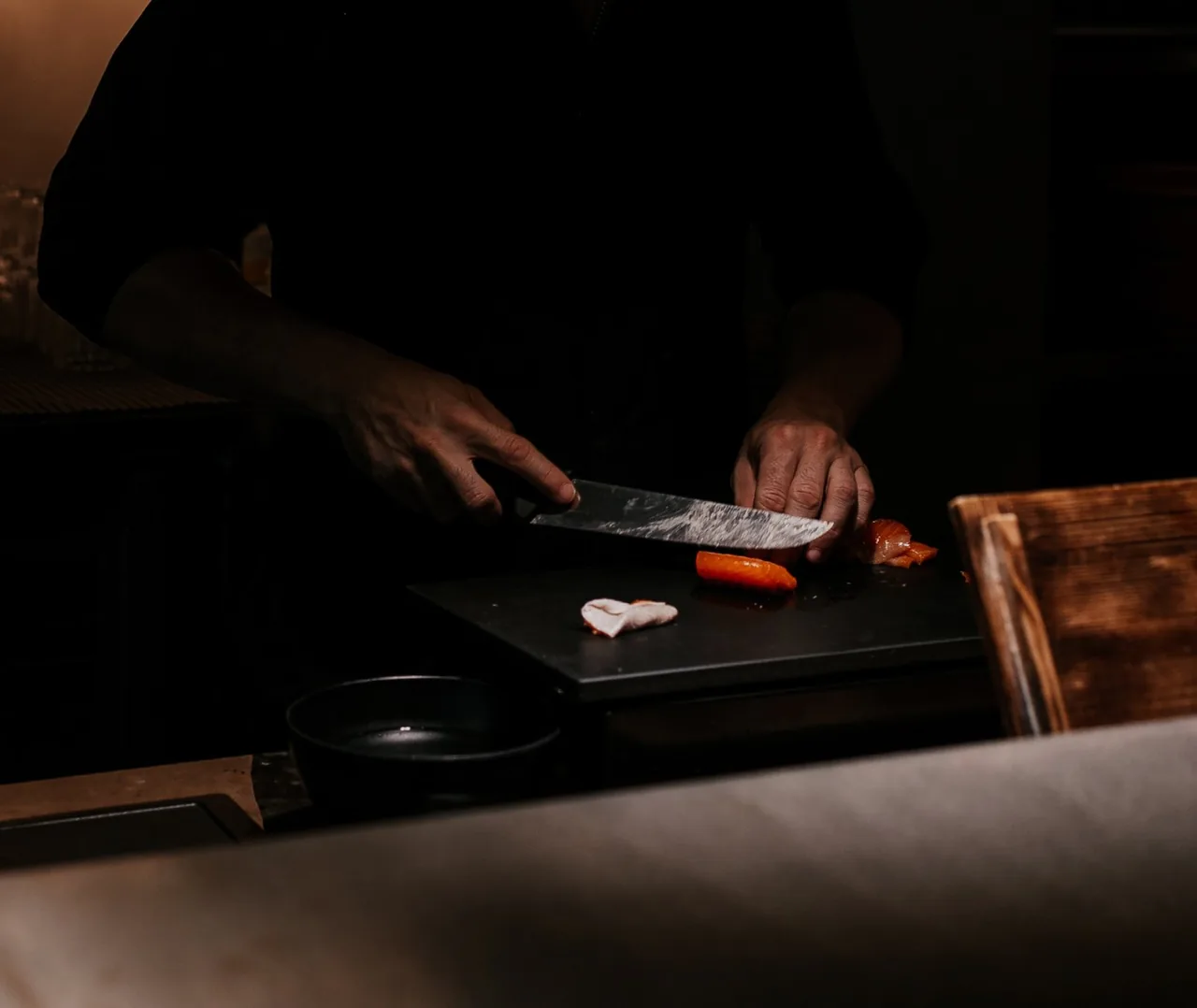 Chef's hands slicing fish with a long knife on a black board at the counter, cut pieces of salmon and white fish beside the blade.