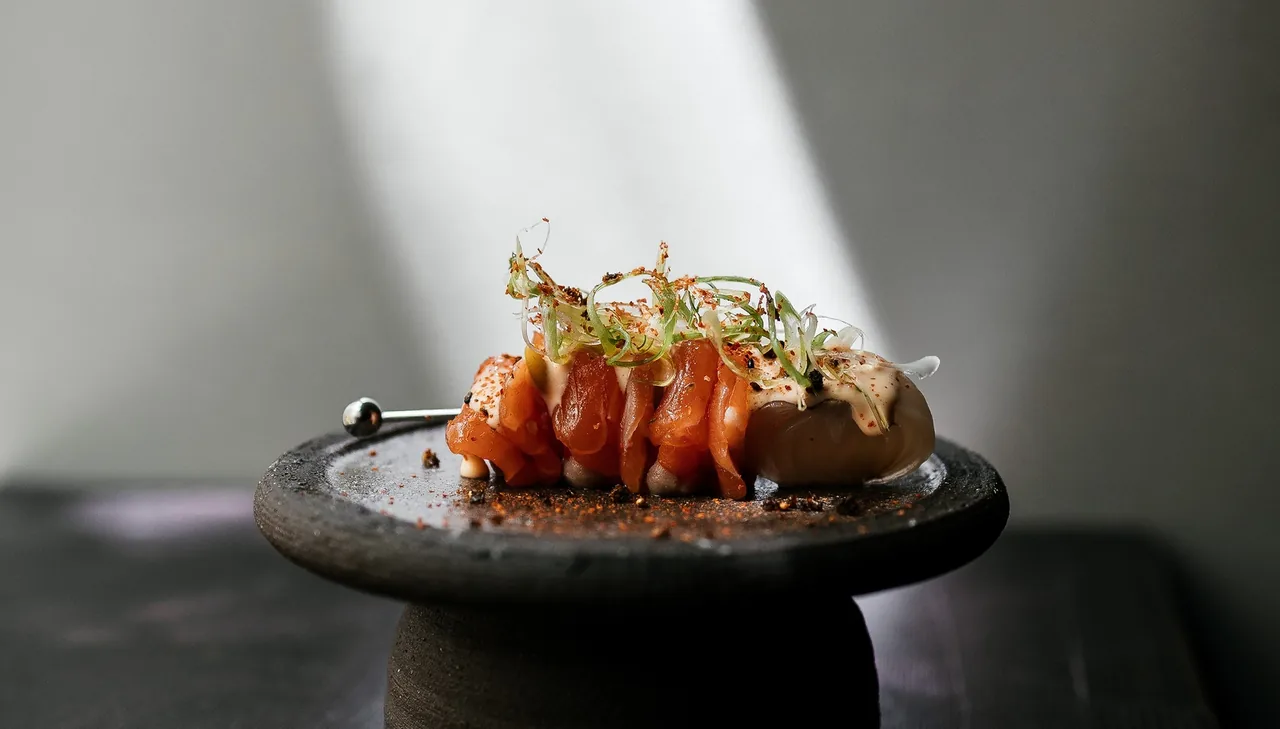 Sliced orange fish topped with fine green shoots and spice on a small dark pedestal plate