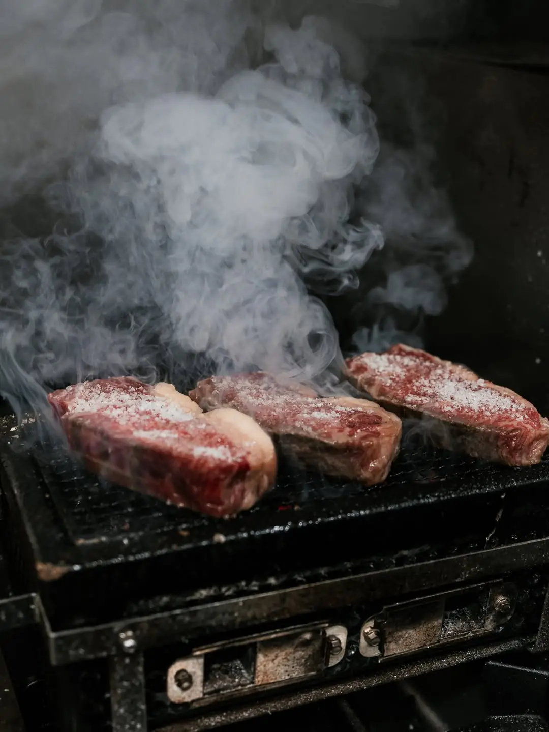 Three seasoned steaks smoking on a charcoal grill