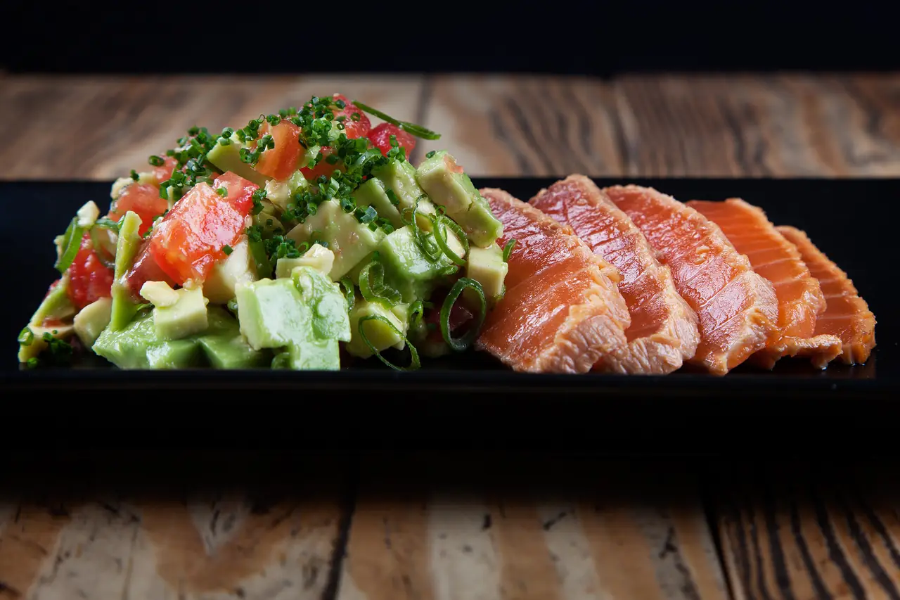 Seared salmon tataki slices beside a diced avocado, tomato, and chive salad on a black rectangular plate, Bambarol