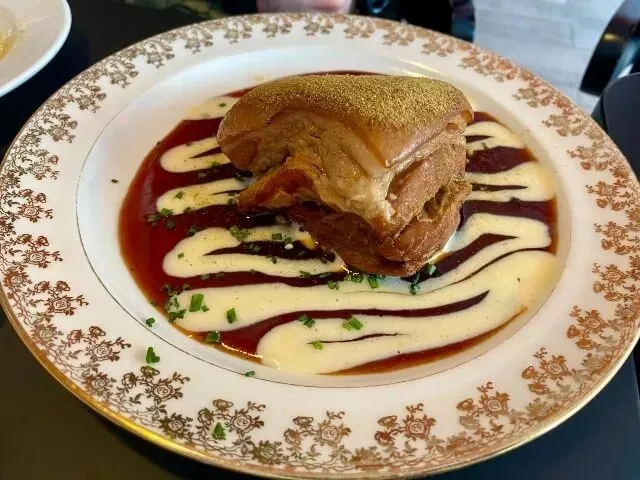 A block of braised pork belly with crisped fat dusted gold, set on dark sauce with piped swirls of pale cream and chopped chives, on a gilt-patterned vintage plate