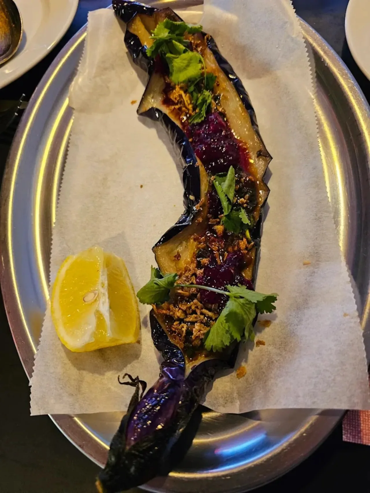 A long charred aubergine split open on greaseproof paper on a steel plate, dressed with dark sauce, crimson puree, crisp fried shallots and coriander leaves, a lemon wedge alongside