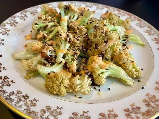 Roasted broccoli and cauliflower florets on a gilt-patterned vintage plate, drizzled with orange sauce and scattered with black and white sesame seeds