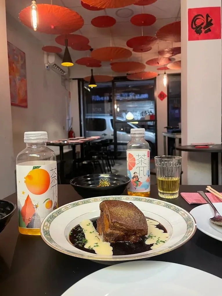 A plate of braised pork belly in dark sauce with pale cream swirls on a green-rimmed plate, two bottles of Chinese fruit tea and a glass of tea behind it, the parasol-covered ceiling of the dining room beyond