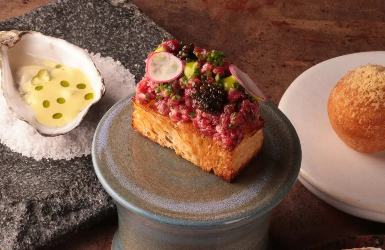 Steak tartare topped with caviar and radish on a slab of toasted brioche, with an oyster in sauce and a croqueta alongside
