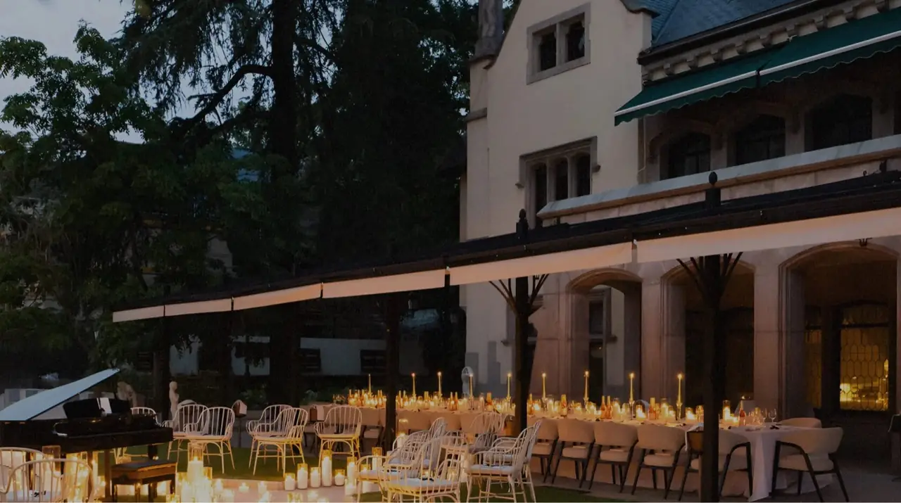 Candlelit outdoor terrace with long white-linen banquet tables and a grand piano beside a historic mansion at dusk
